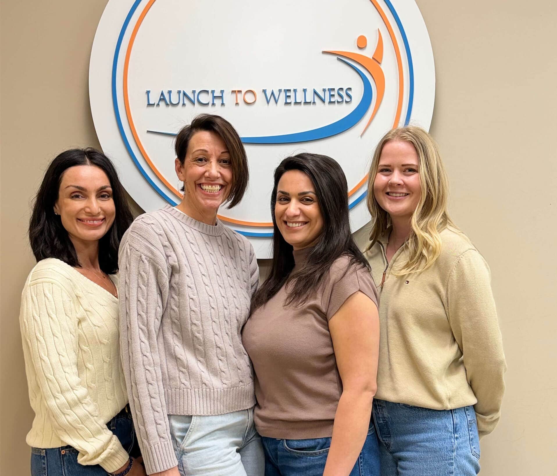 Four team members smiling in front of the Launch To Wellness wall logo