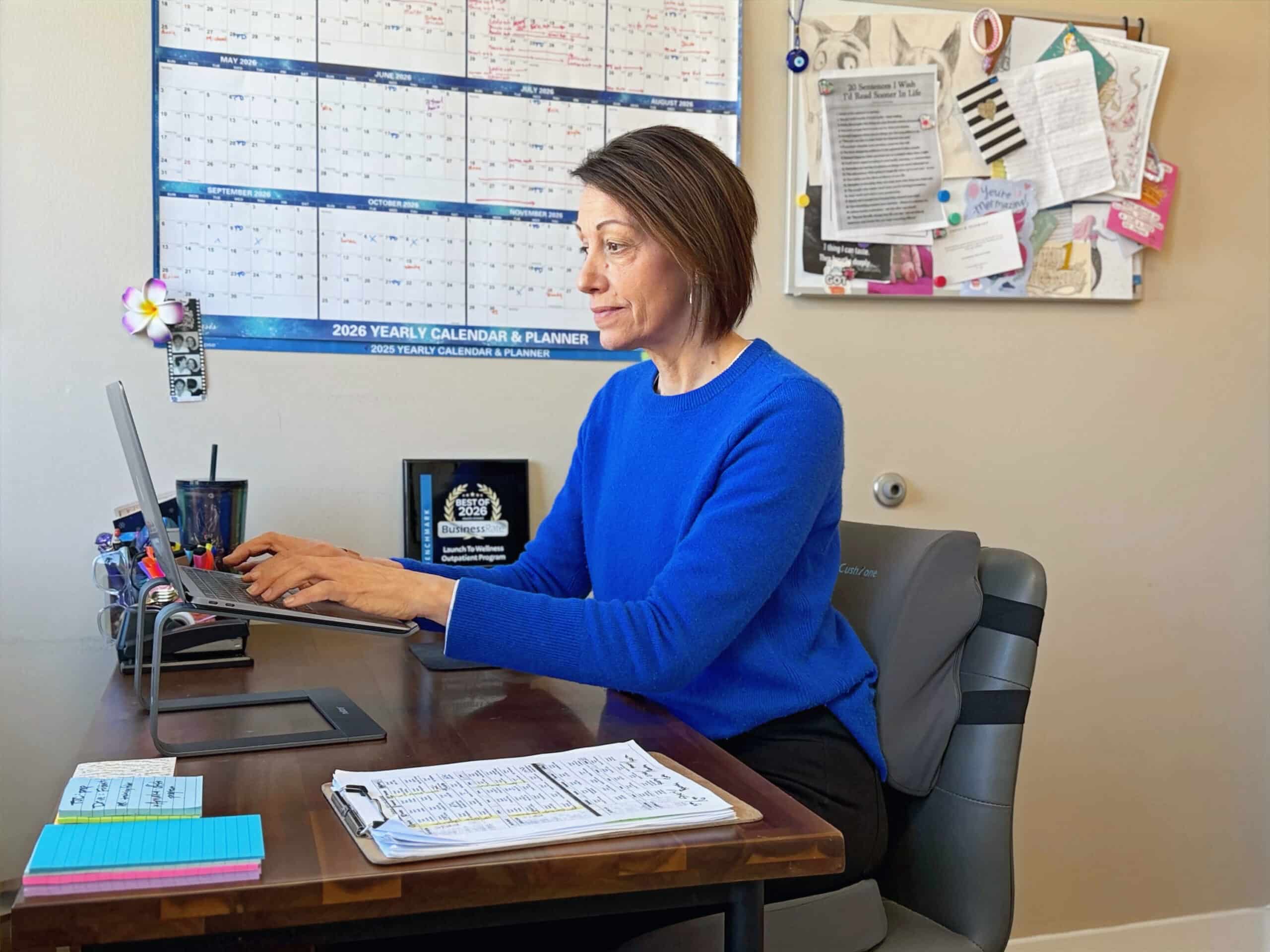 Clinician at a desk with a laptop, yearly calendar, and session notes
