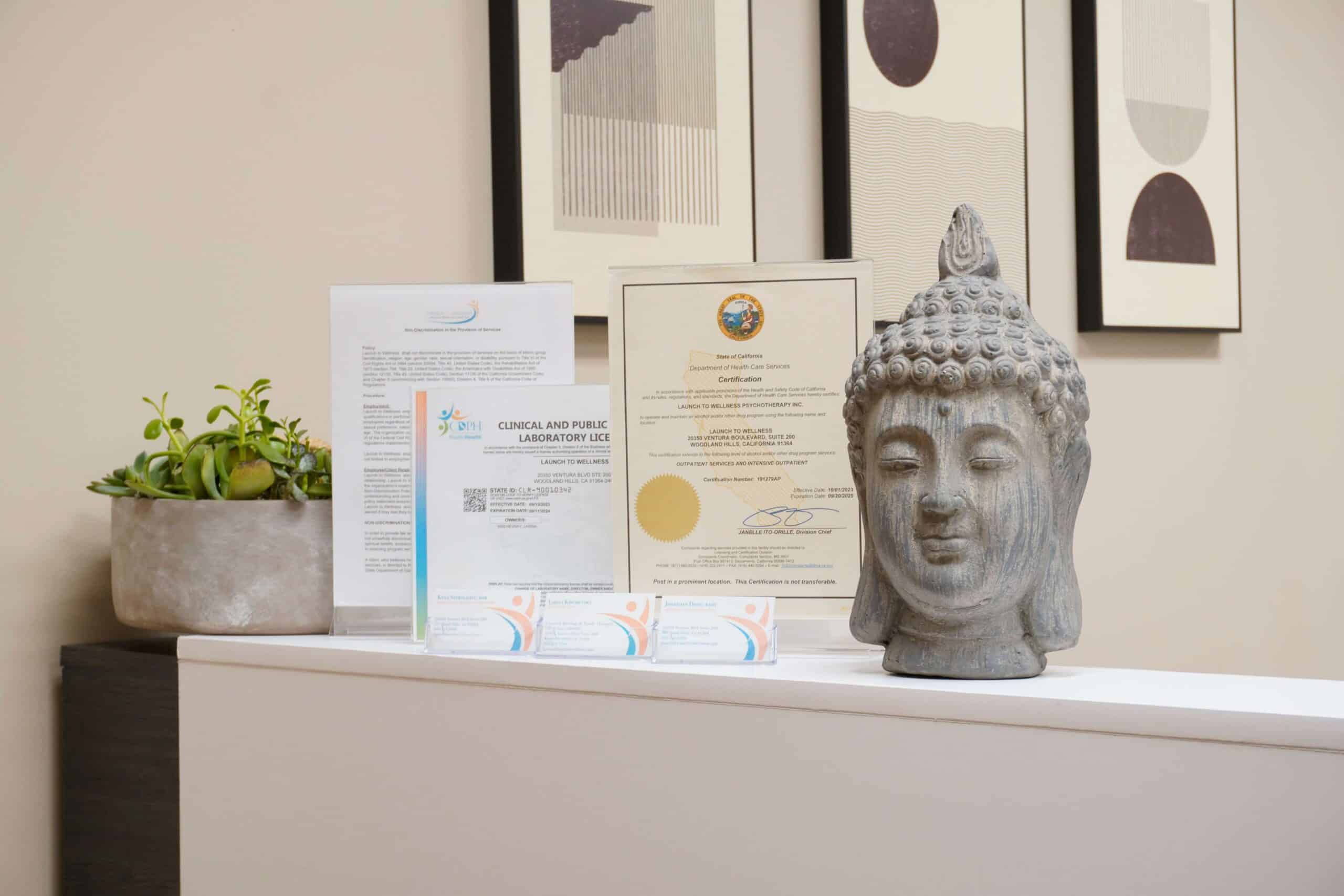 A stone Buddha head beside California treatment certifications on a shelf at Launch To Wellness.