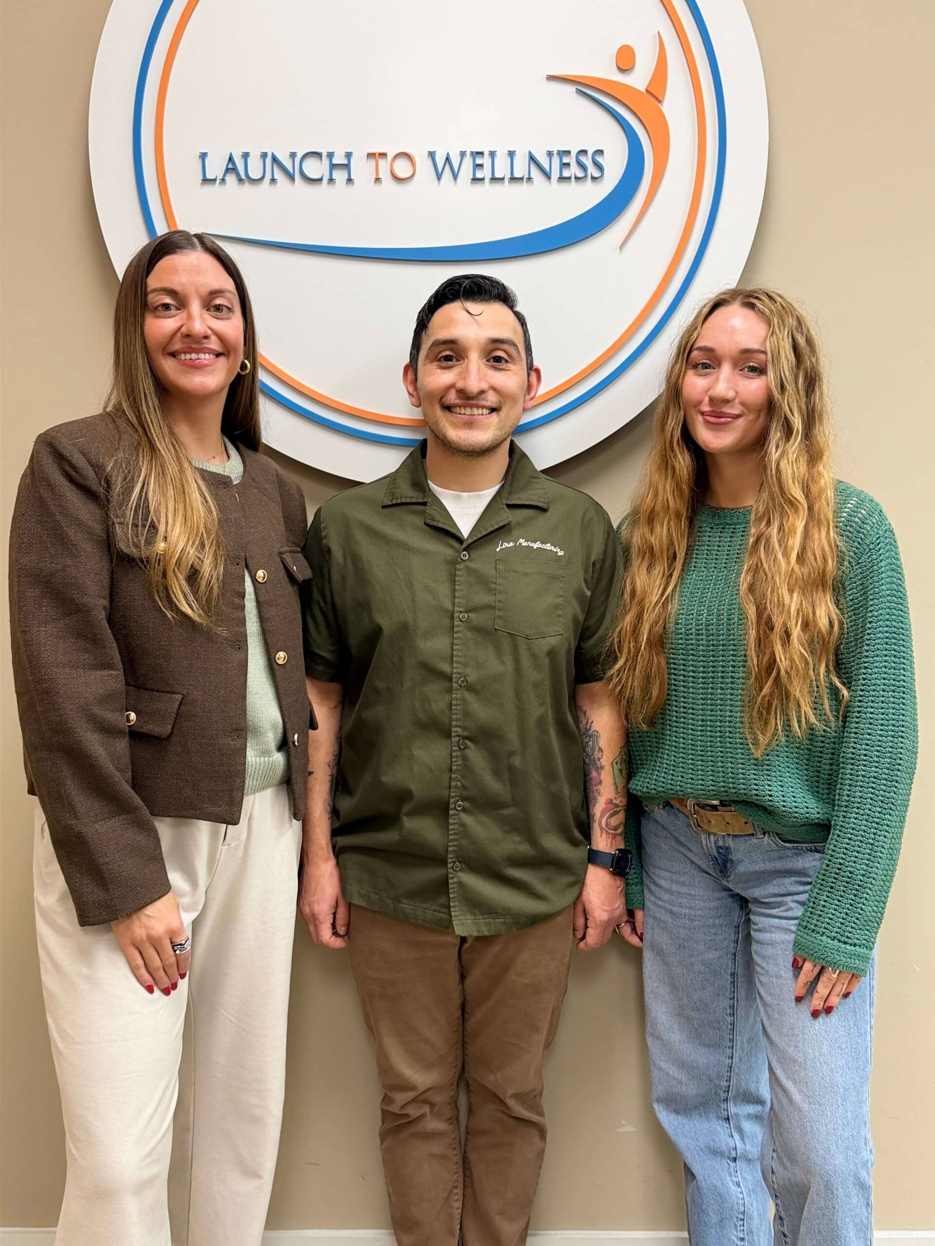 Three team members standing in front of the Launch To Wellness wall logo