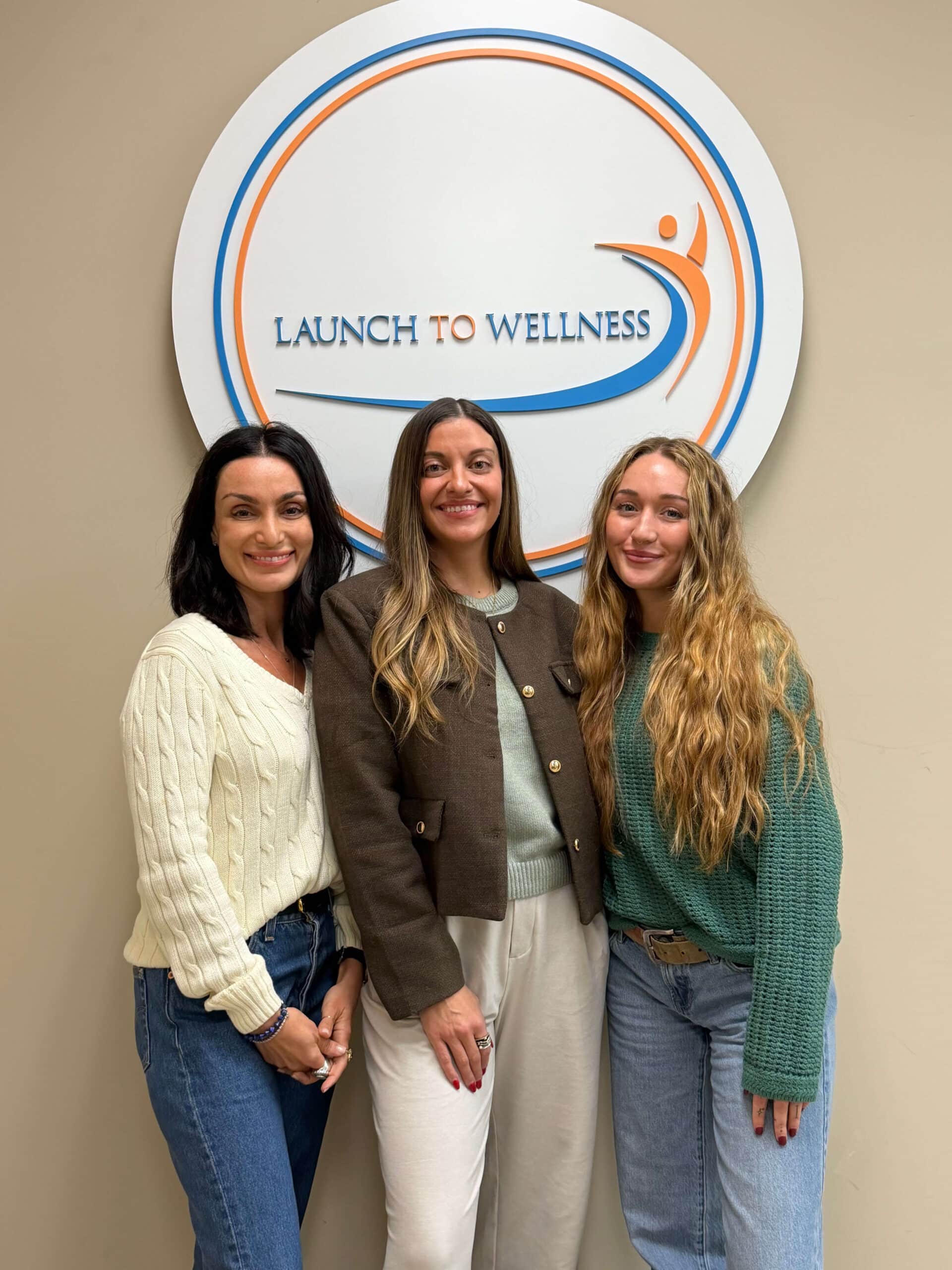 Three team members smiling in front of the Launch To Wellness wall logo